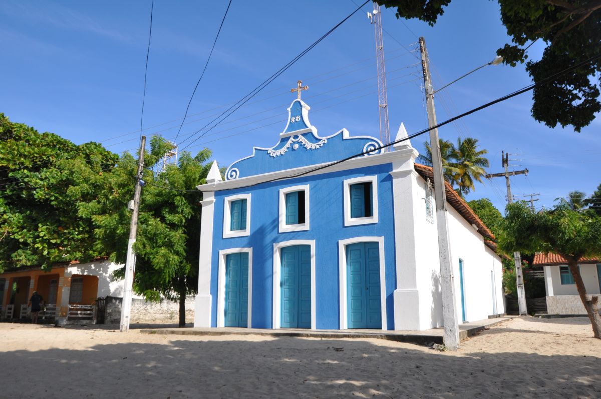 Pequena casa azul em cima do mangue, rodeada por plantas e com o céu nublado.